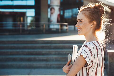 Cheerful caucasian girl with red hair and freckles is holding a tablet and looking away staying in front of a building